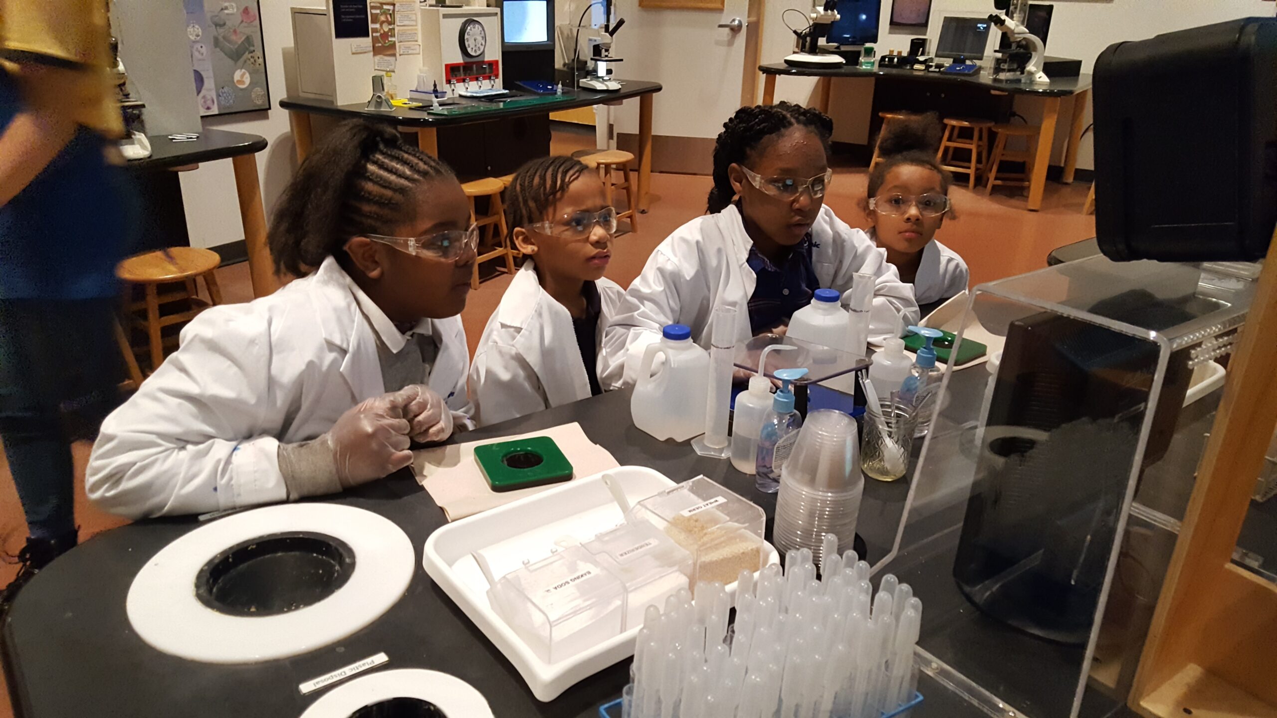 Four students wearing lab coats and goggles conducting an experiment in a science classroom