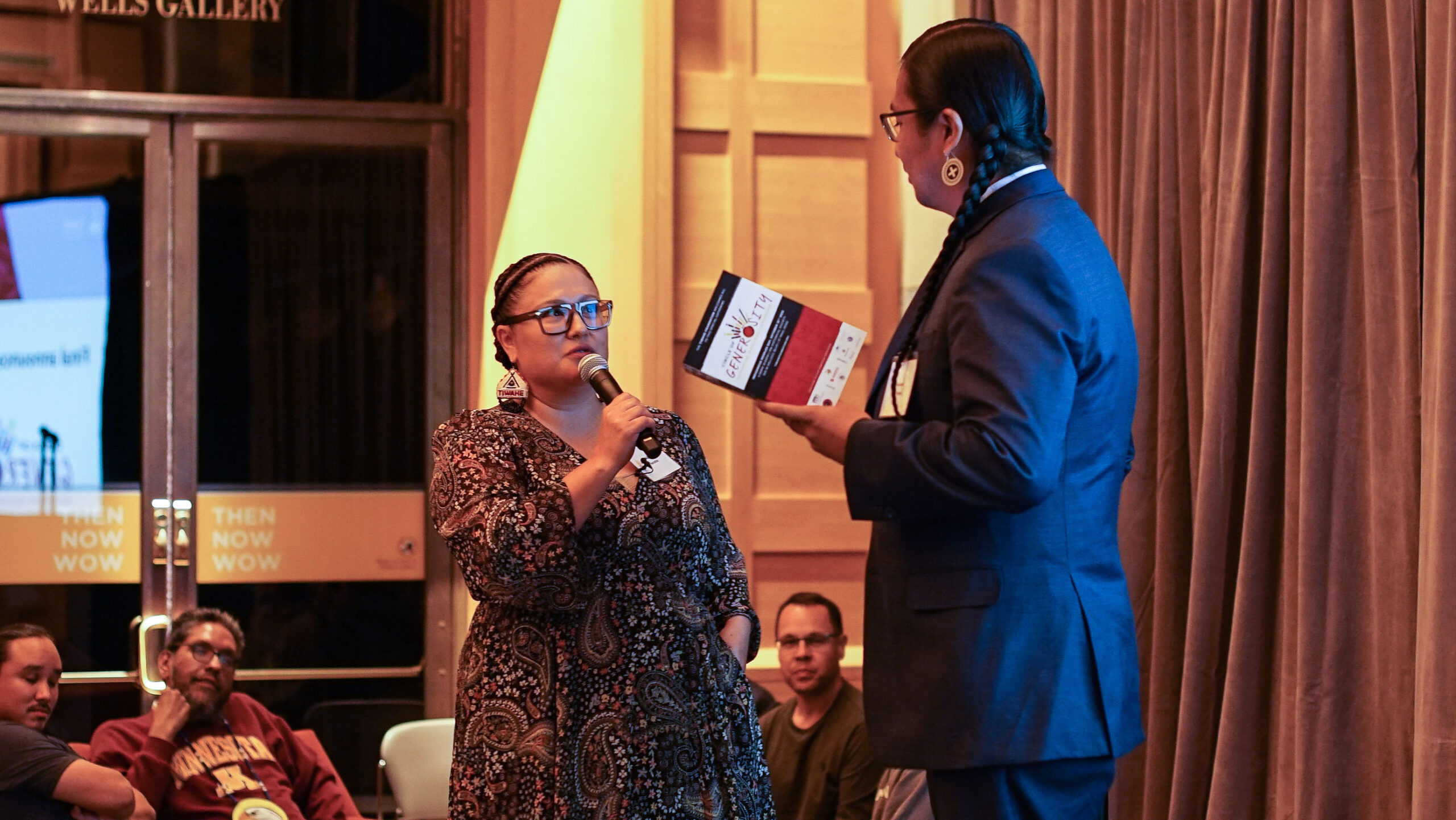 Person on stage with microphone speaks to another person holding book